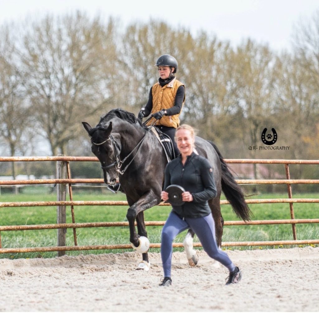 Ruiter traint op de grond haar enkelstabiliteit terwijl een andere ruiter voorbij komt gereden op een zwart paard.